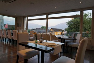 Un comedor con una mesa dispuesta para dos, ventanas amplias que ofrecen vistas a montañas y casas, y una fila de sillas blancas.