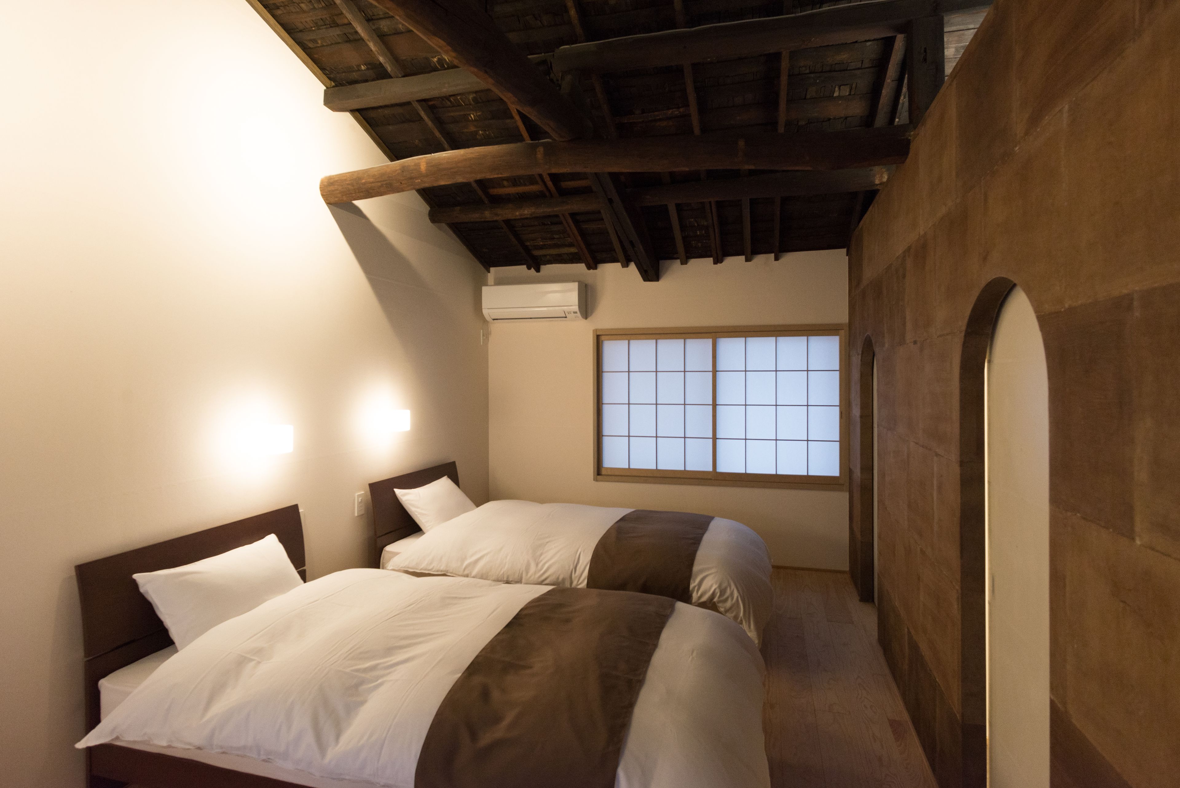 A room with two beds, a wooden ceiling, a window, and an air conditioning unit.