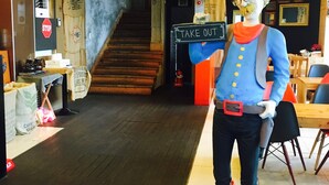 Een standbeeld van een personage in uniform met laarzen, staand op een platform in een kamer met houten vloer, een trap en een 'TAKE OUT' bord.