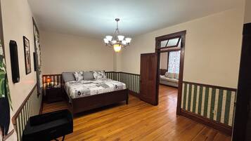 Ein Schlafzimmer mit Holzboden, einem Bett mit gemustertem Überwurf, einem Sessel, einem Schreibtisch, einer Kristallleuchtre und einer Tür zu einem anderen Raum.