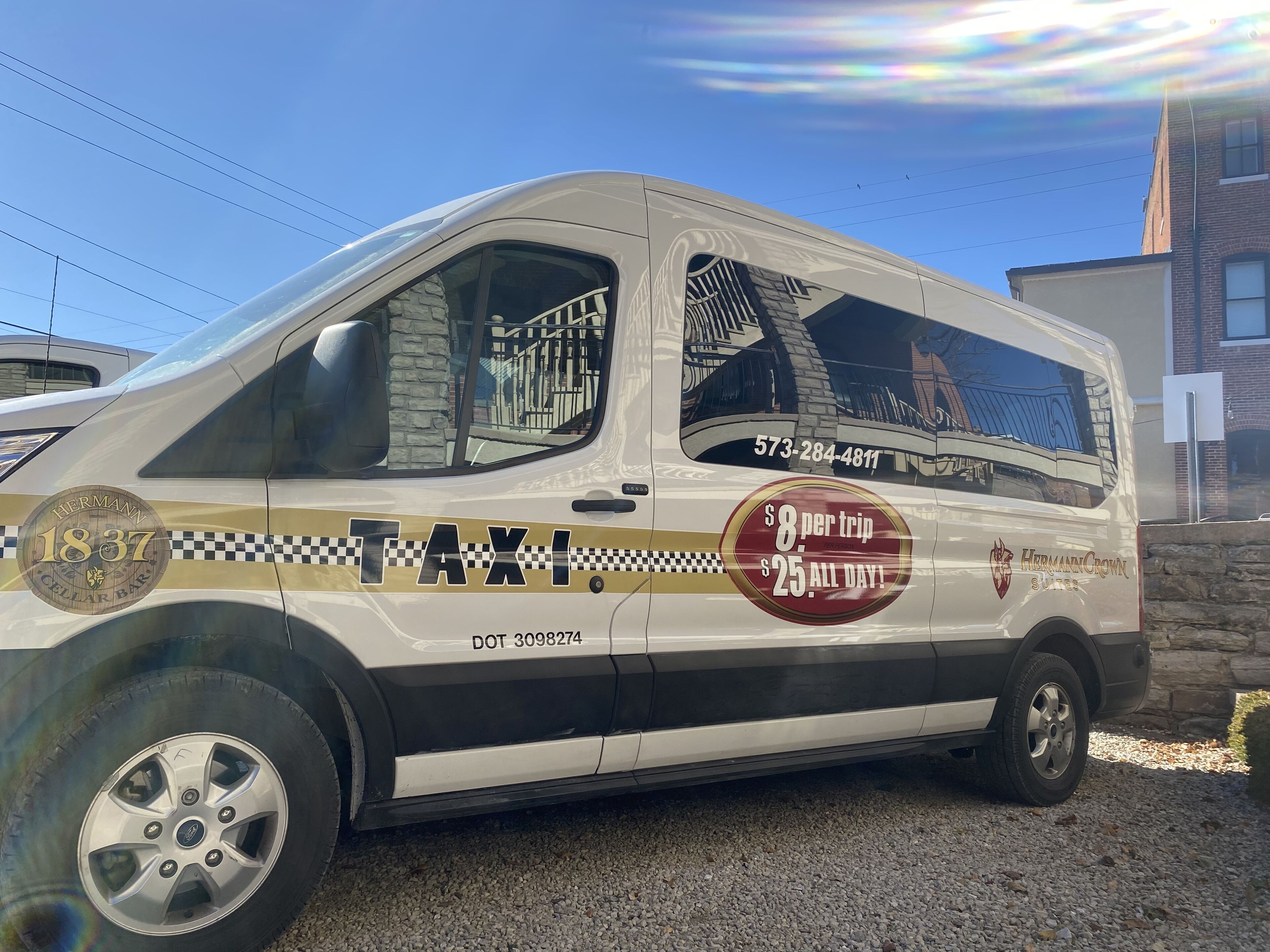 Una camioneta blanca con la leyenda "Hermann 1837" y la palabra "TAXI", estacionada sobre una superficie de grava.