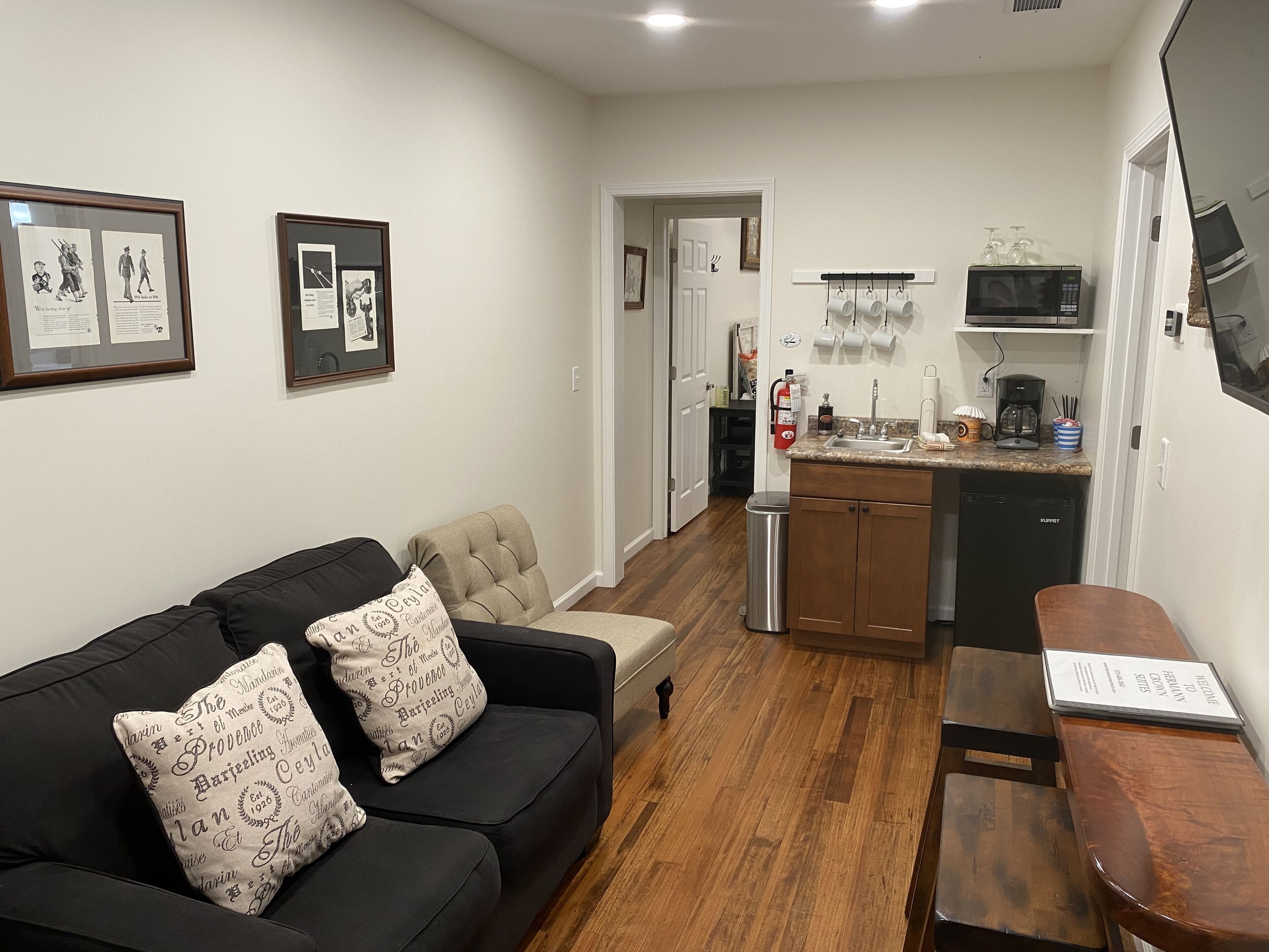 A living room with a black sofa, a beige armchair, and a wooden bench. There are framed pictures on the wall, a kitchen area with a microwave, sink, and cabinets, and a fire extinguisher on the wall.