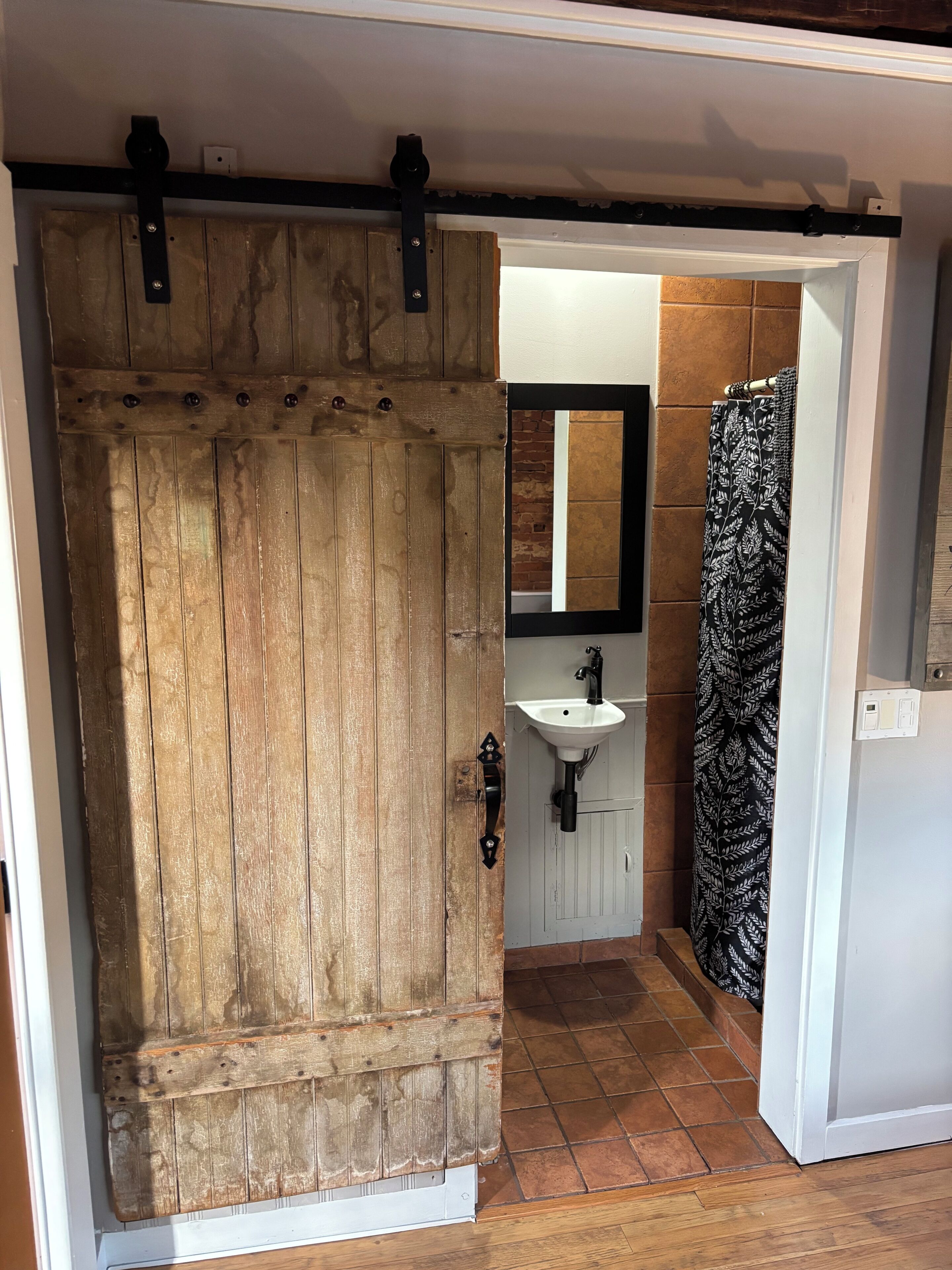 A rustic wooden door with a black metal handle, a bathroom with a white sink and mirror, and a shower with a black and white patterned curtain.