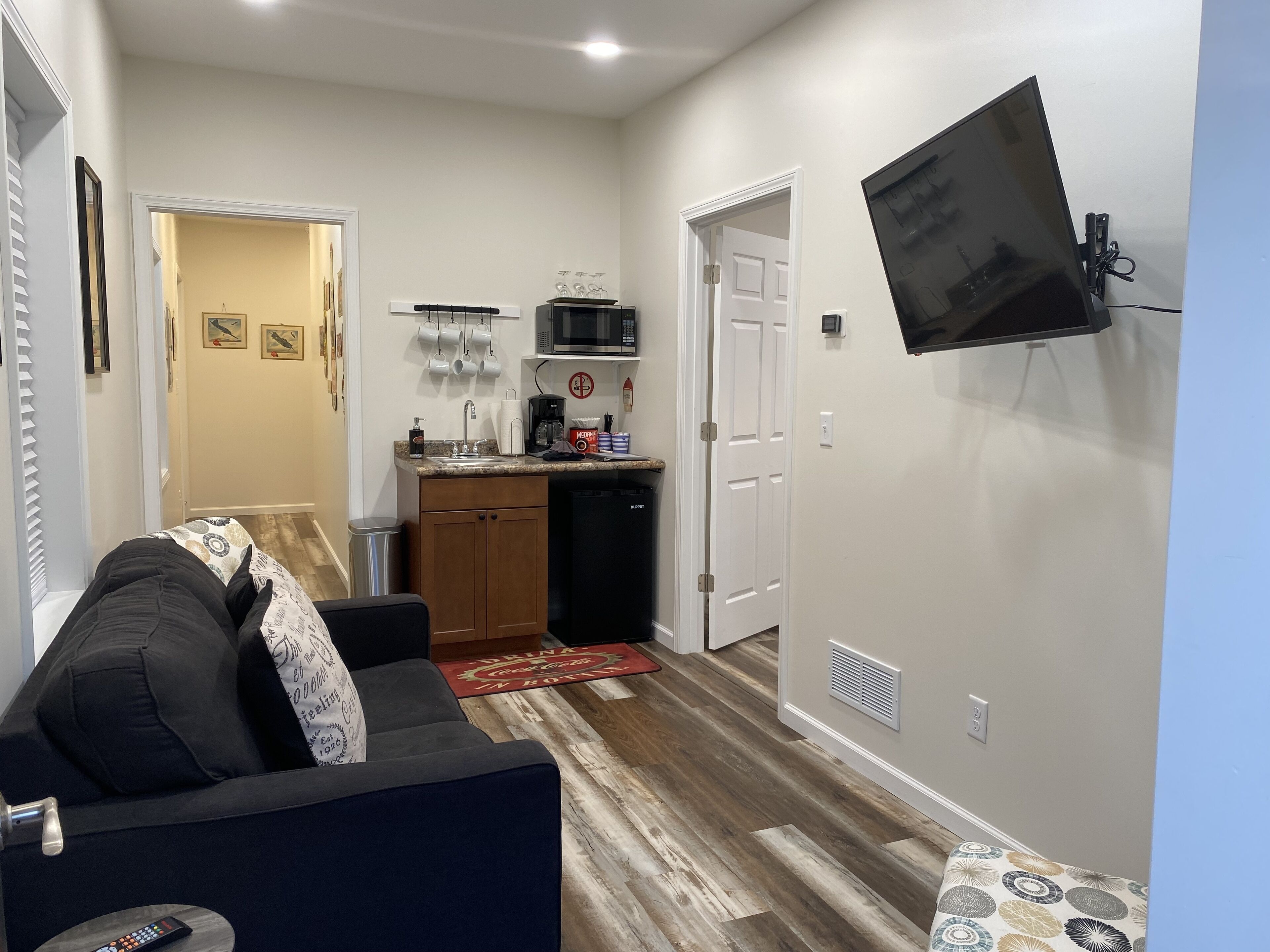 A compact living space with a sofa, a flat-screen TV mounted on the wall, a kitchenette with a microwave, and a coffee maker.