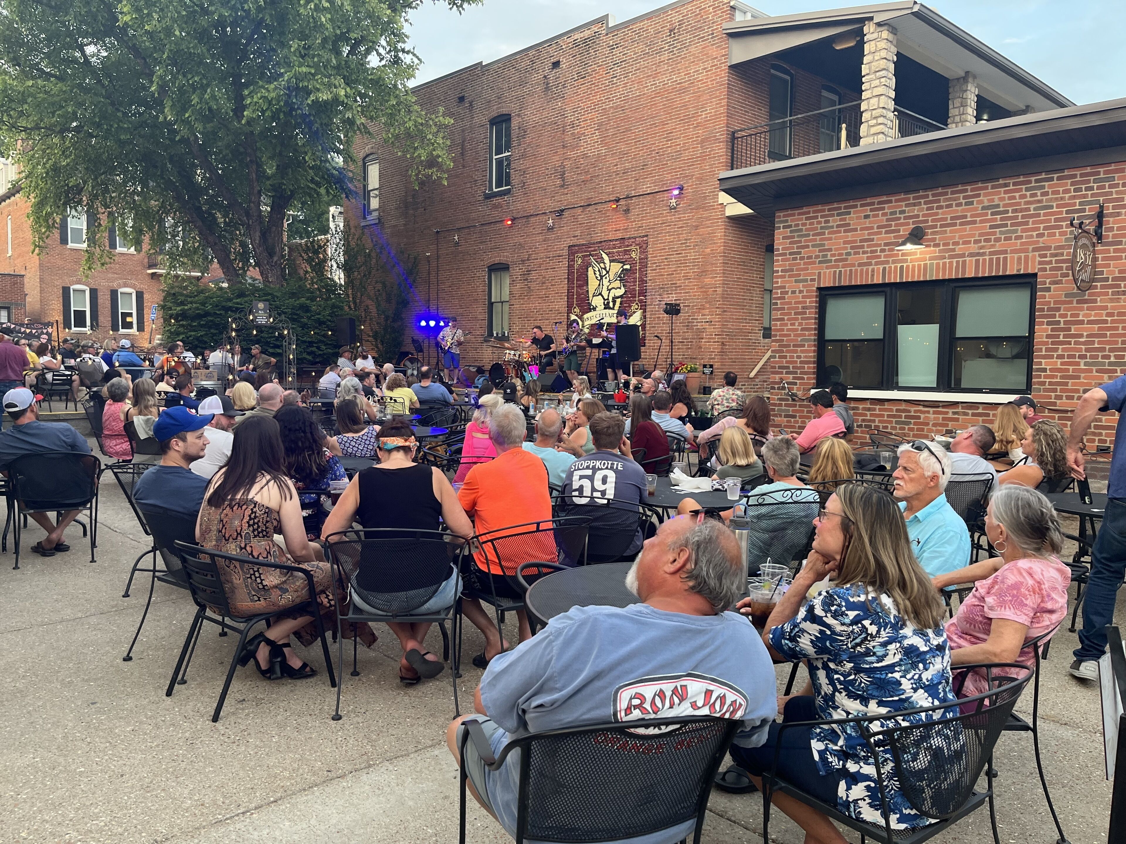 An outdoor gathering with people seated at tables, a stage with musicians, and a brick building in the background.