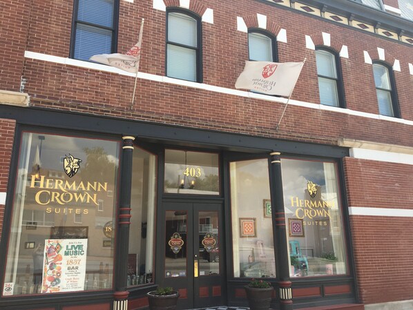 A building with large windows displaying the name "Hermann Crown Suites". There are flags on the roof, and a sign for live music events is visible on the window.