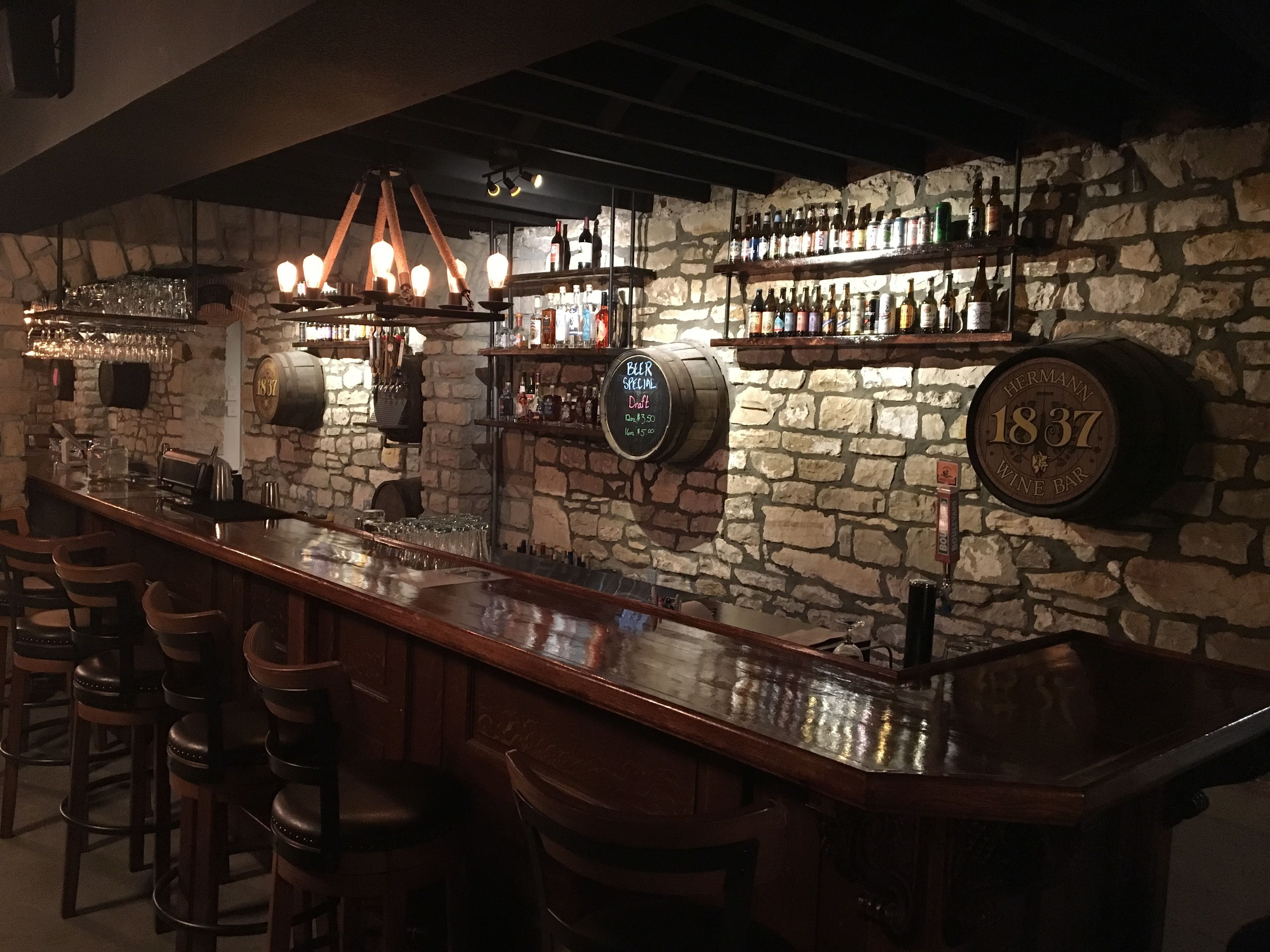 A bar with stone walls, wooden bar stools, and a variety of bottles and glasses on shelves.
