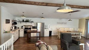 A spacious living area with a sofa, armchair, and dining table. A kitchen with white cabinetry and a 'Butter & Eggs' sign is visible. - Hermann Crown Suites (Hermann)