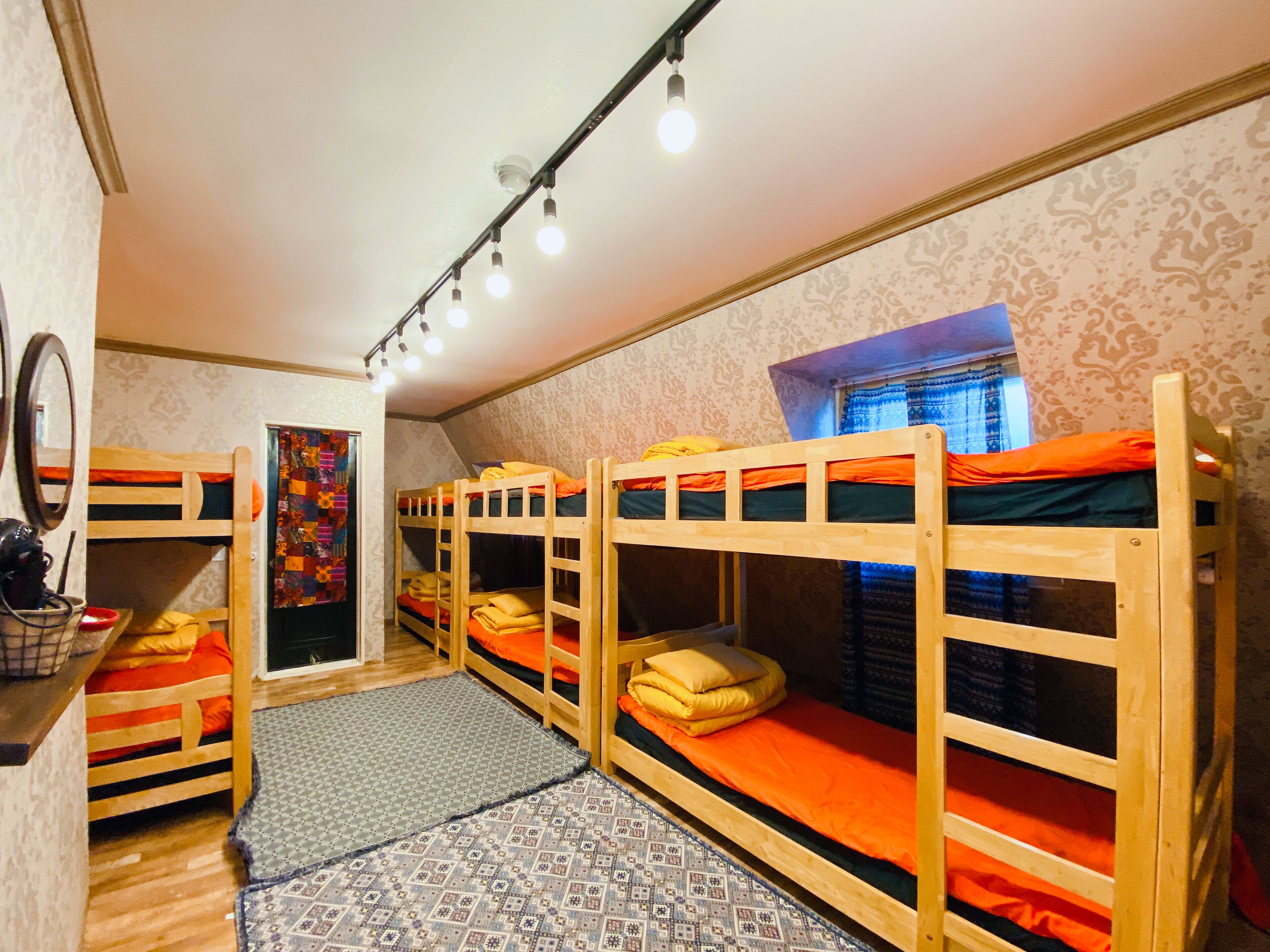 A dormitory room with bunk beds, a mirror, a patterned rug, and a window with curtains.