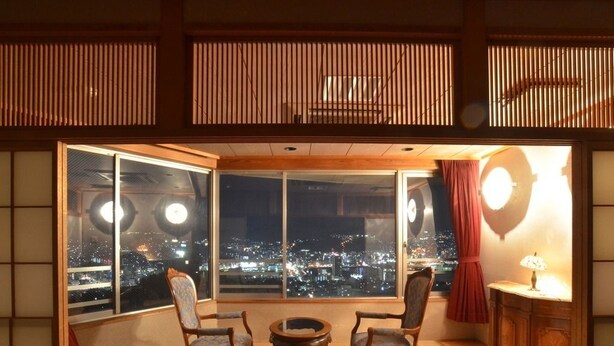 Une pièce dotée d’une grande fenêtre donnant sur la ville de nuit, d’une porte coulissante de style japonais traditionnel et d’une petite table avec des chaises.