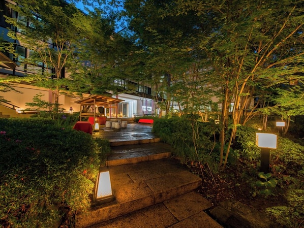 A well-lit outdoor area with steps leading to a covered seating area, surrounded by lush greenery and trees.