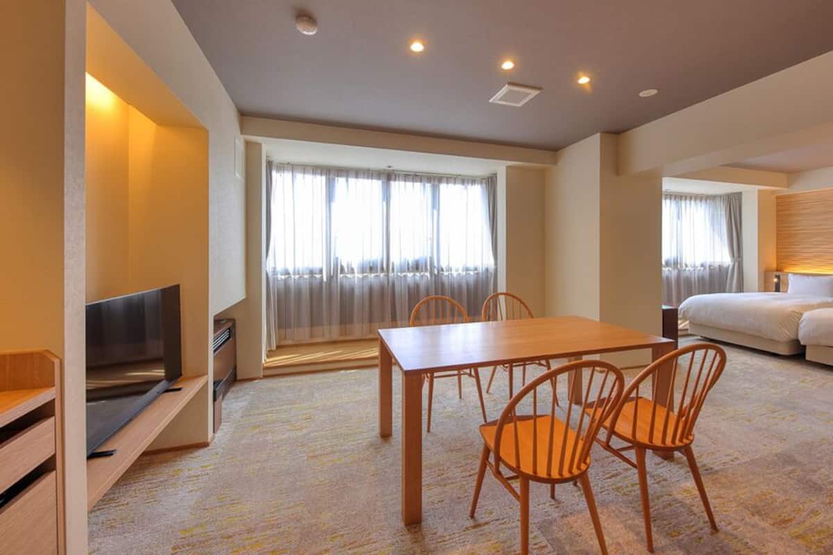 Habitación de hotel con televisor, una mesa de comedor con sillas, sofá y un amplio ventanal con cortinas.