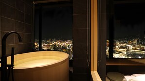 Ein modernes Badezimmer mit einer großen Badewanne, einem Fenster mit Blick auf die Stadt und einem kleinen Nachttisch.