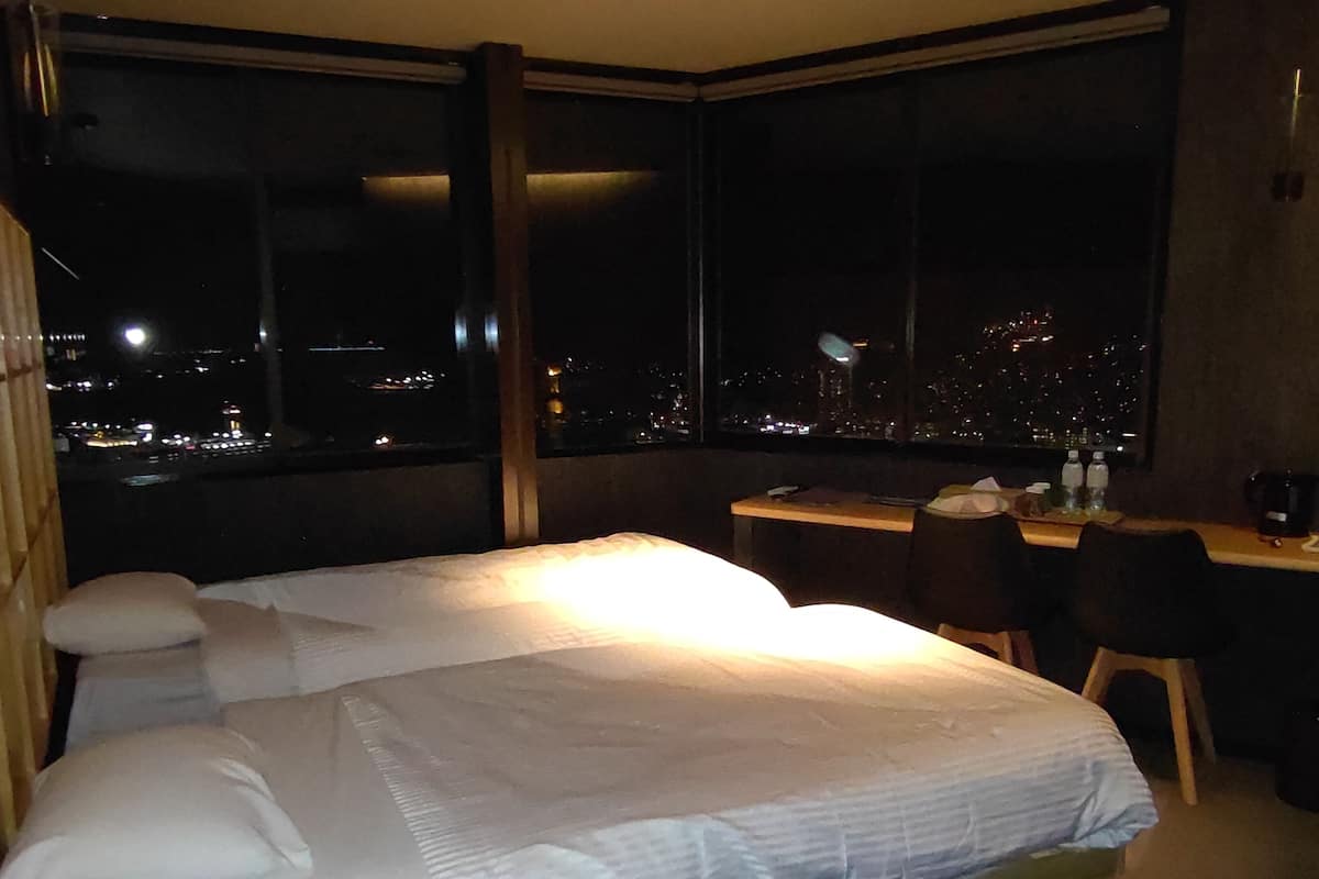 Habitación de hotel con cama, escritorio y silla. Se observa una vista nocturna de la ciudad a través de amplias ventanas.