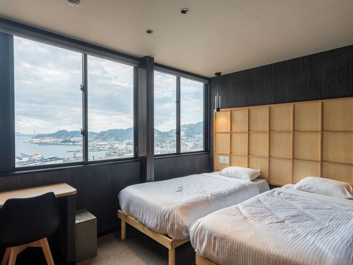 Habitación de hotel con dos camas, un escritorio y un amplio ventanal con vistas a la ciudad y a las montañas.