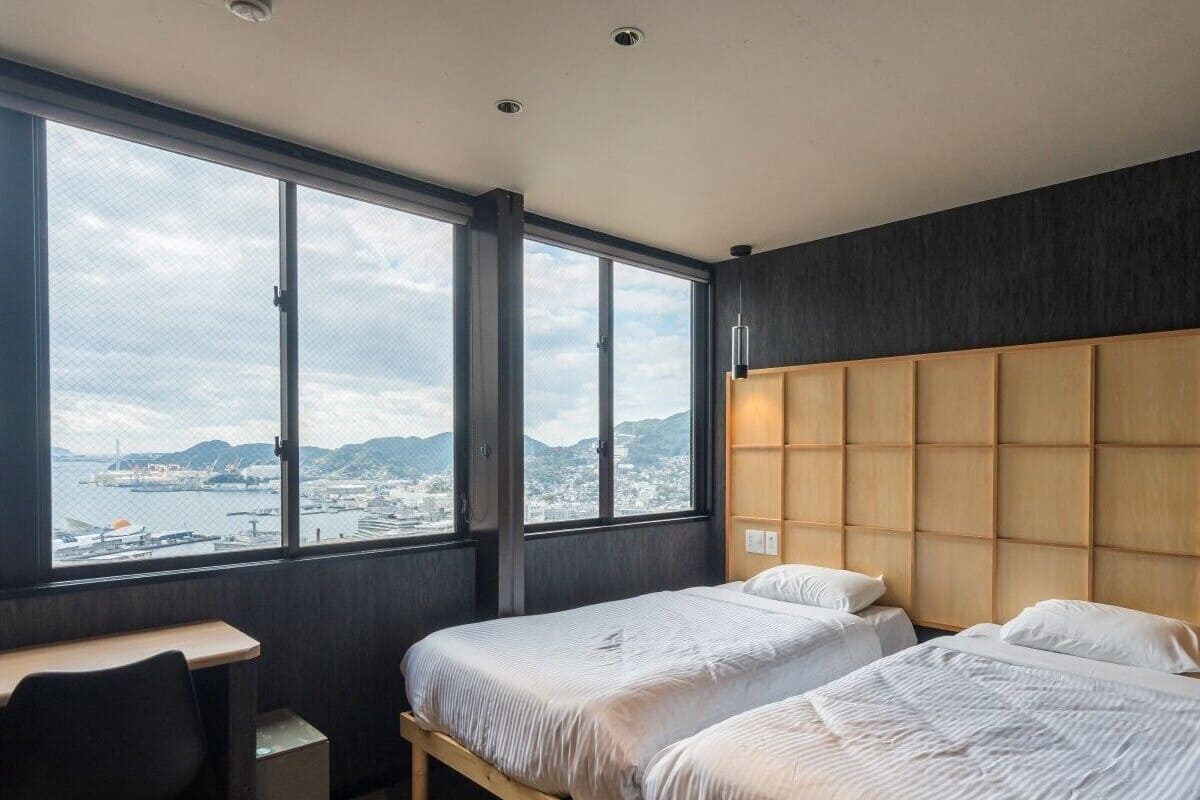 Habitación de hotel con dos camas, un escritorio y un amplio ventanal con vistas a la ciudad y a las montañas.