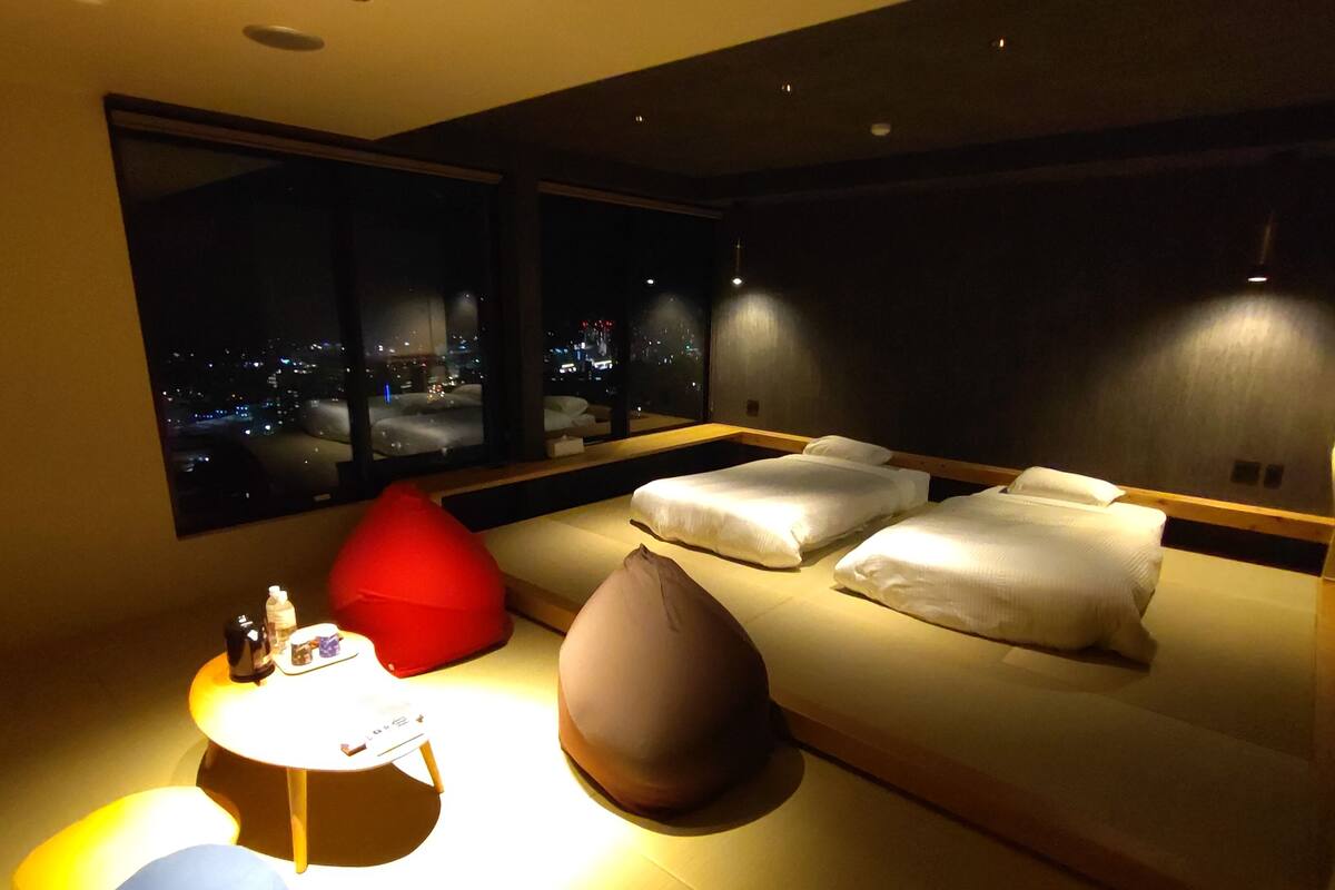 Una habitación de hotel moderna con una cama grande, una mesita redonda pequeña y un sillón de peluche rojo.