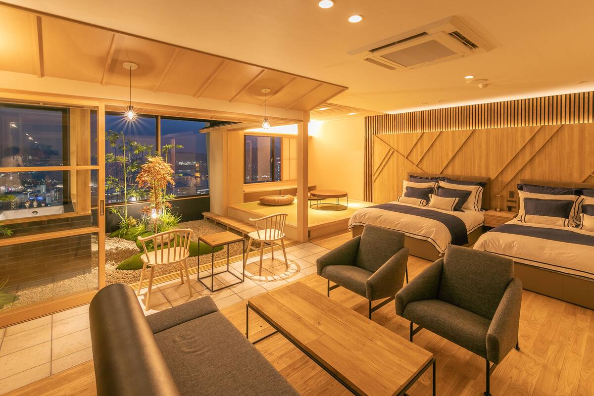 Una habitación de hotel moderna con una cama grande, dos mesitas de noche, un sofá, una mesita pequeña y una silla. Hay un ventanal con vistas a la ciudad de noche.