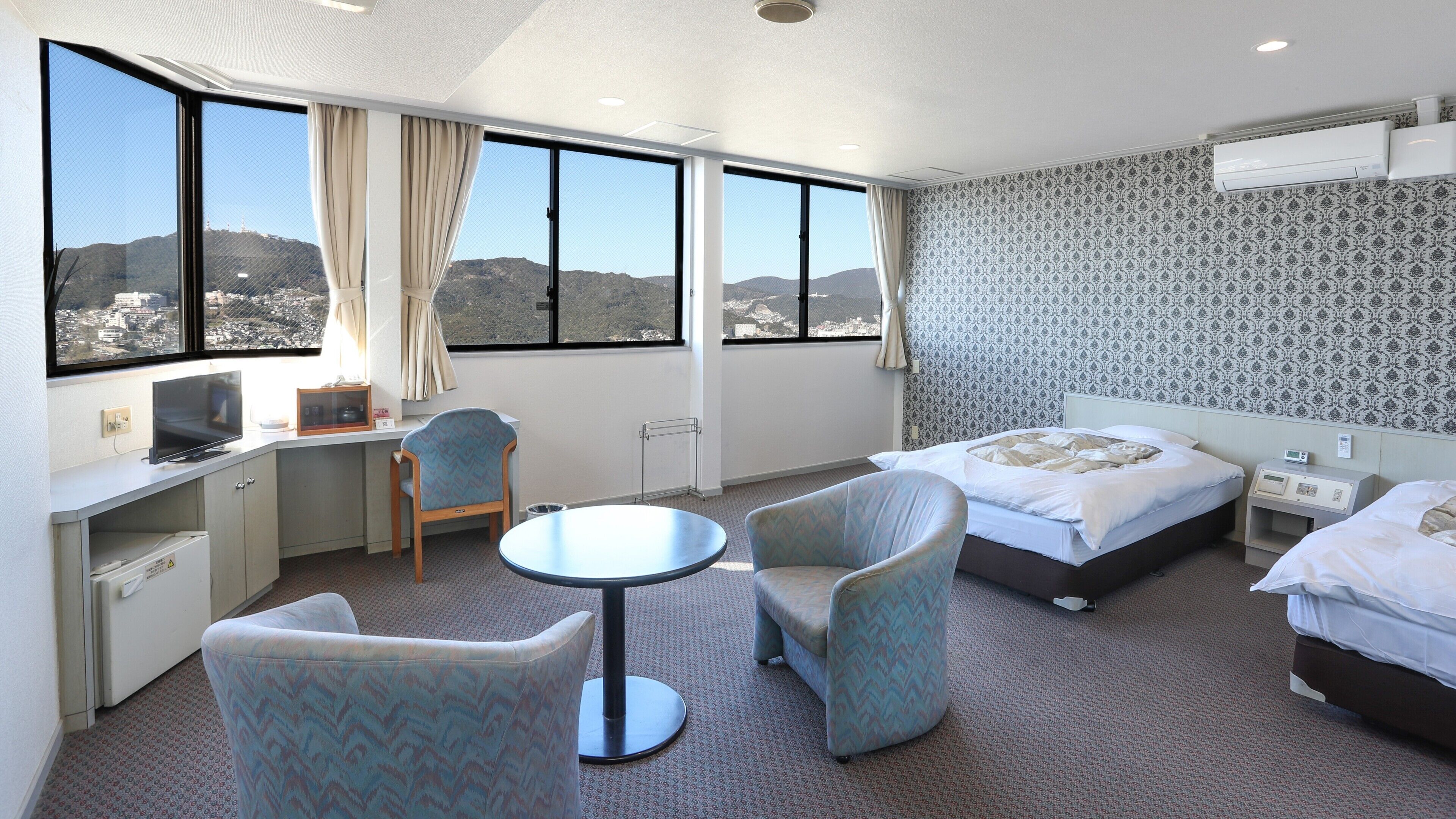 Ein Hotelzimmer mit Schreibtisch, zwei Stühlen, einem Bett und Blick auf eine Stadtlandschaft durch große Fenster.