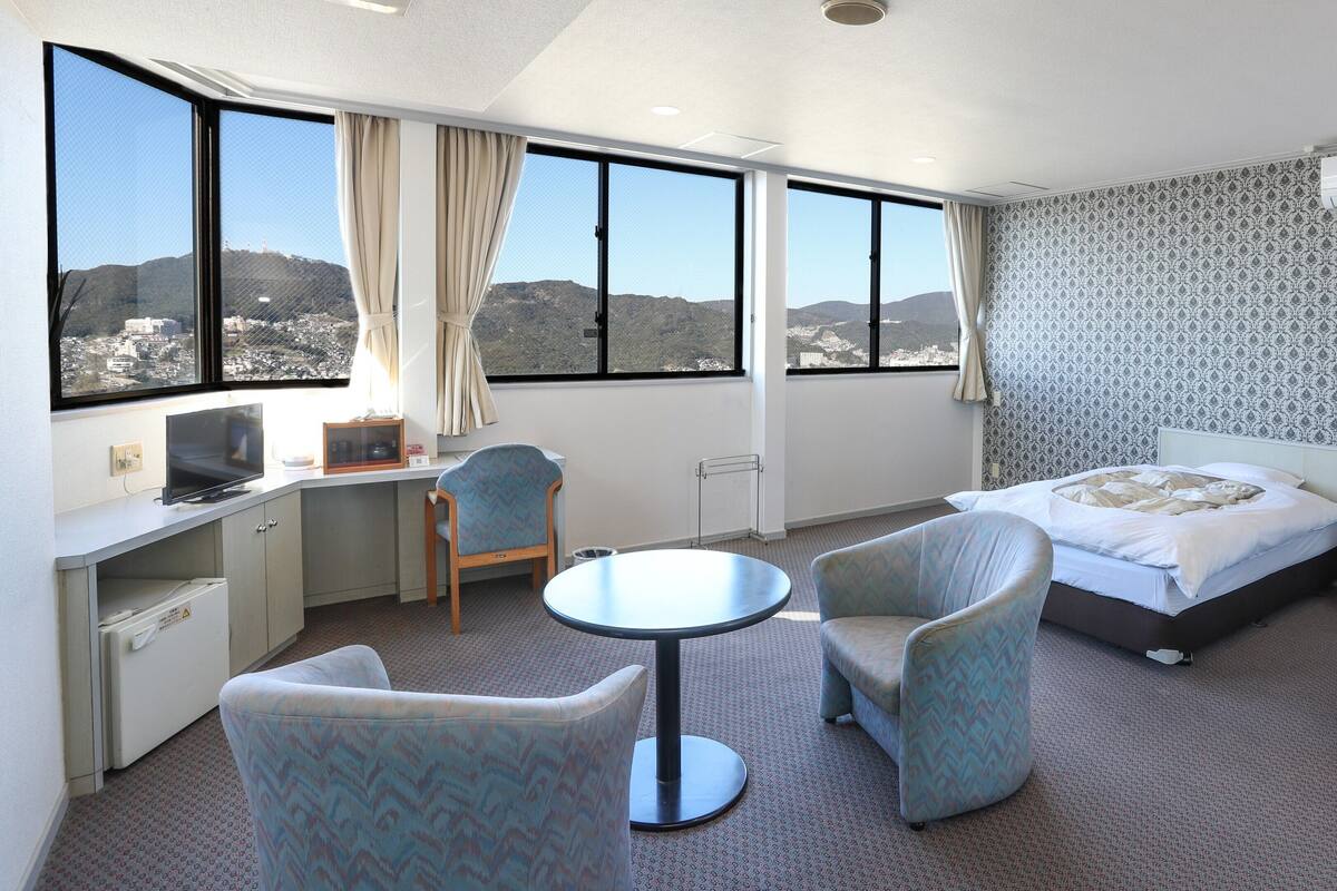 Habitación de hotel con escritorio, dos sillas, una cama y vistas a un paisaje urbano a través de amplios ventanales.
