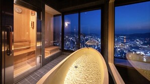 Ein moderner Wellnessraum mit großer Badewanne, Holzsauna und Blick auf die Stadt im Abenddämmerung.