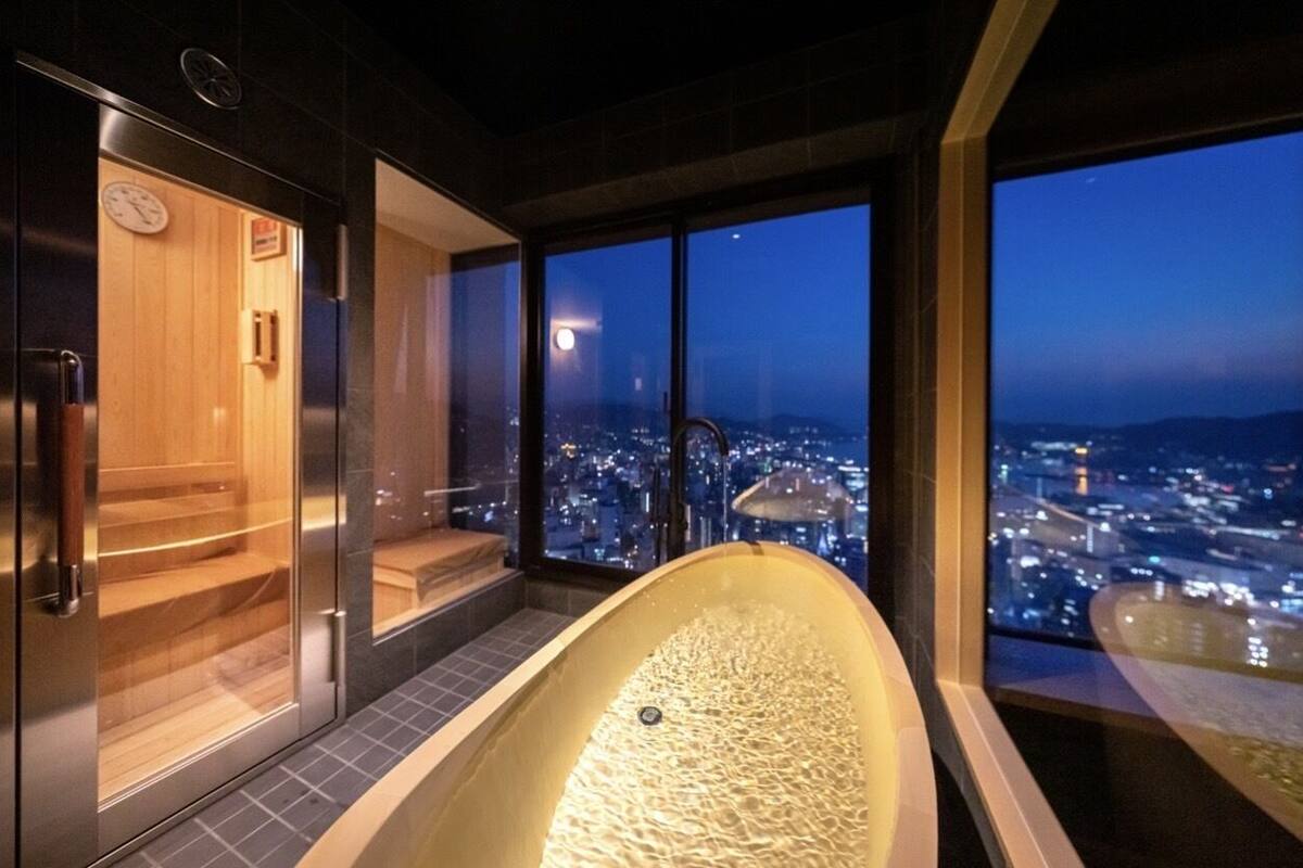 Un moderno spa con una bañera grande, sauna de madera y vistas a la ciudad al anochecer.