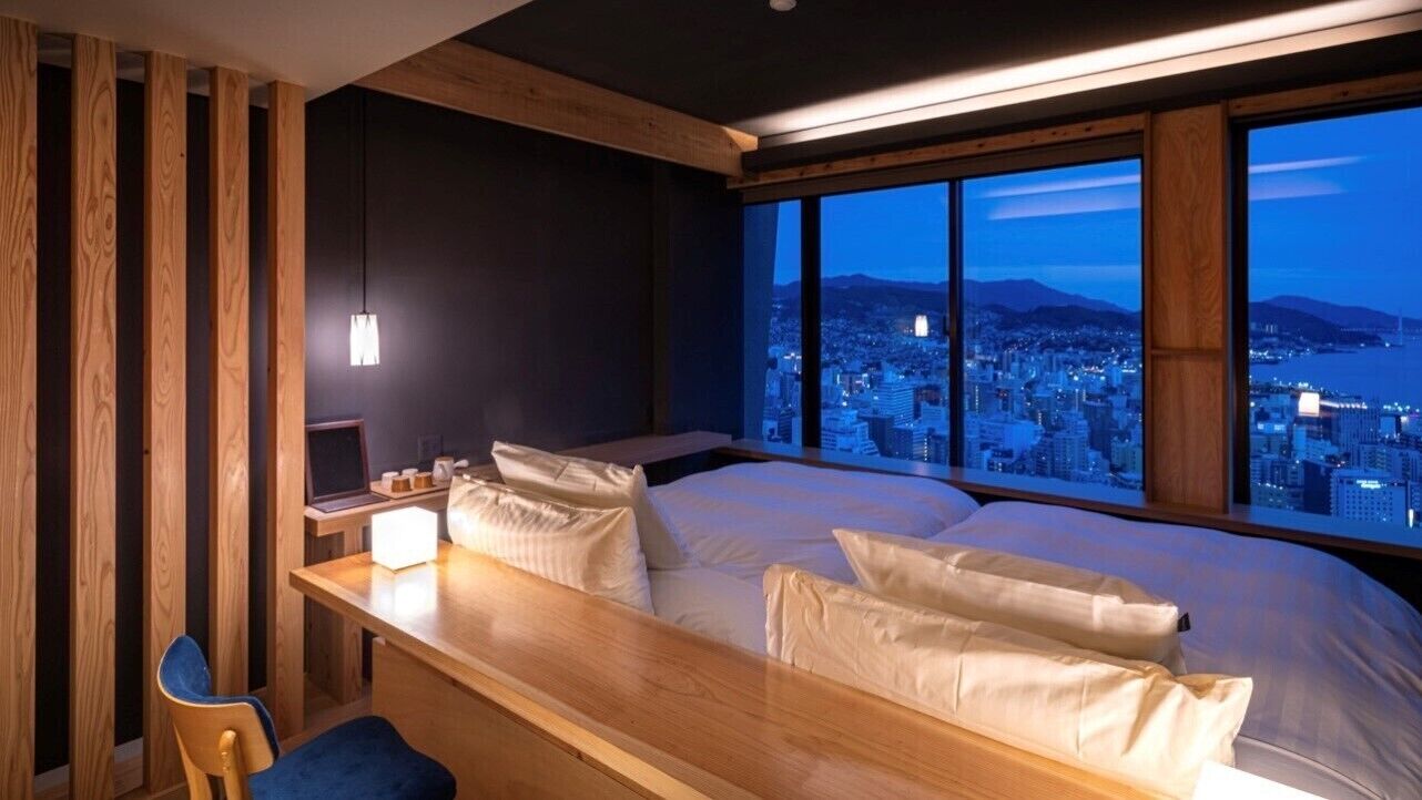 Ein Schlafzimmer mit einem großen Bett, einem Schreibtisch und Blick auf eine Stadtlandschaft im Abenddämmerung.