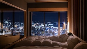 Ein Schlafzimmer mit großem Fenster und Blick auf eine beleuchtete Stadt bei Nacht.