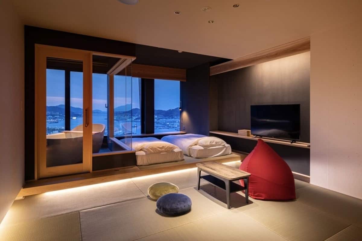 Una sala moderna con un ventanal grande, un televisor de pantalla plana y un área de descanso con cojines y un sillón de peluche rojo.