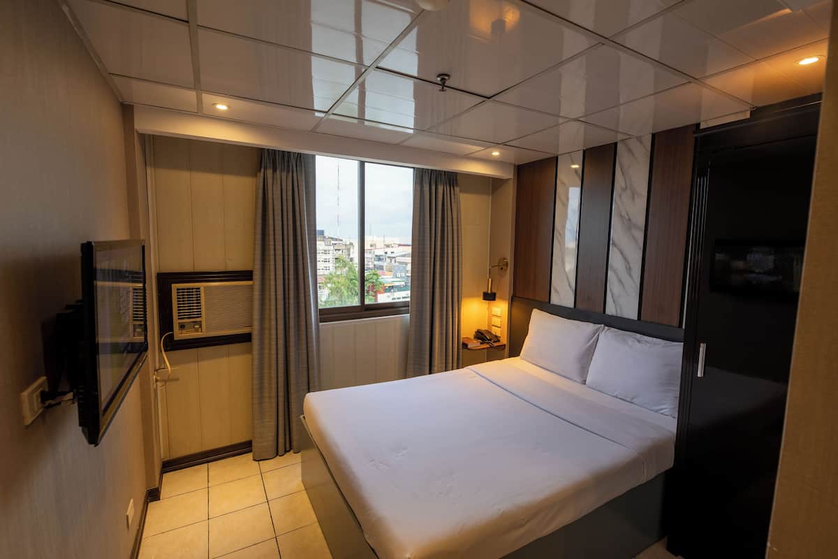 Habitación de hotel con cama, televisor, ventana con cortinas, mesita de noche con lámpara y vistas a la ciudad.