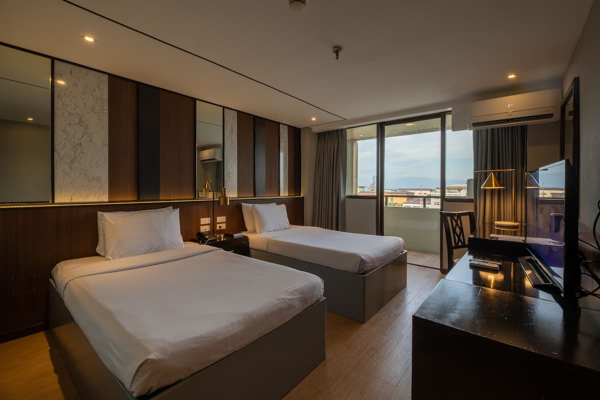 Habitación de hotel con dos camas, un escritorio con televisor y un ventanal con vista a la ciudad.