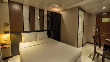 A hotel room with a large bed, a desk with a chair, a wardrobe, and a bedside lamp.