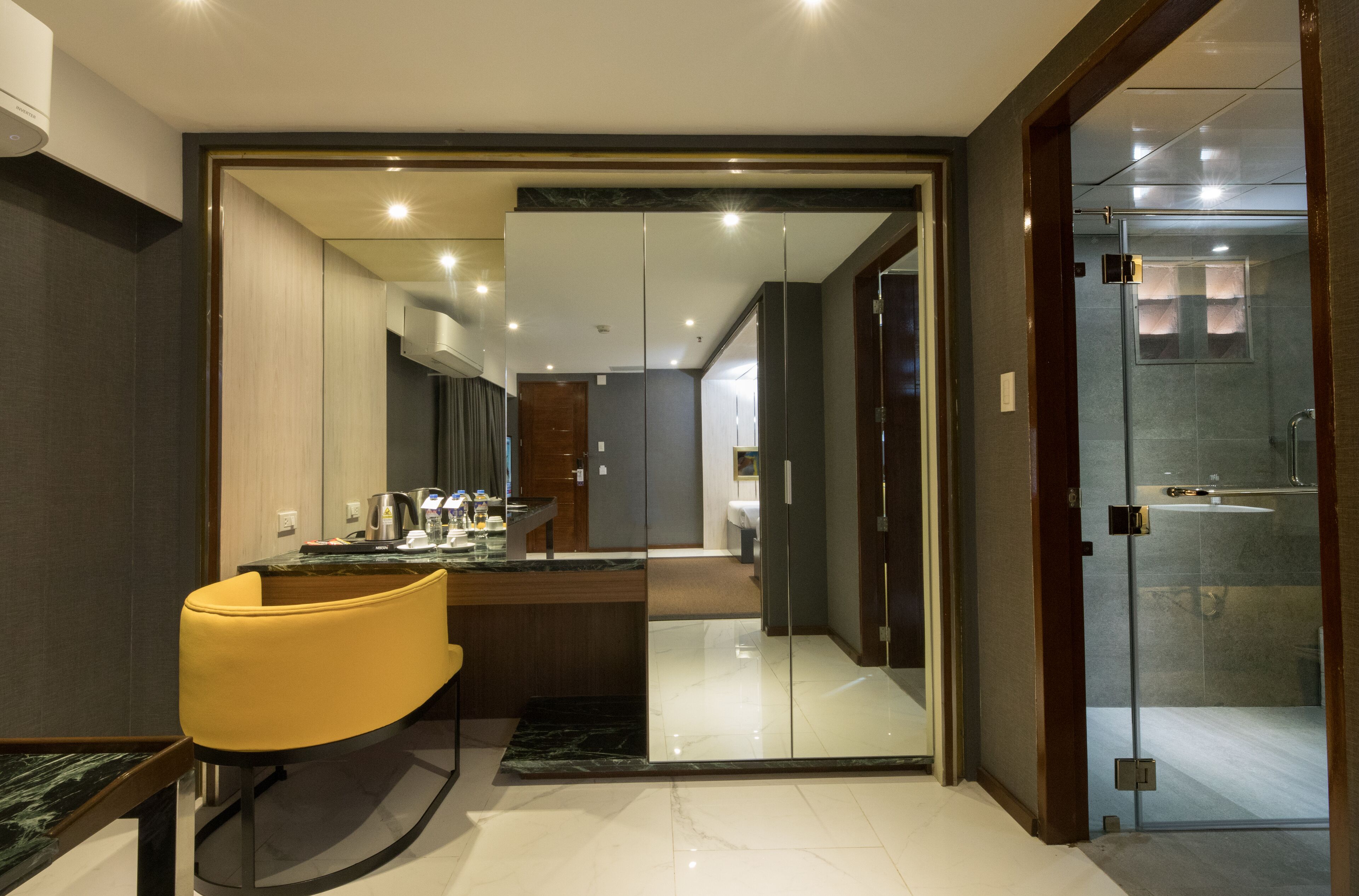 Une chambre d’hôtel moderne dotée d’une douche à parois de verre, d’un coin repas avec une chaise jaune et d’une cuisine ouverte.
