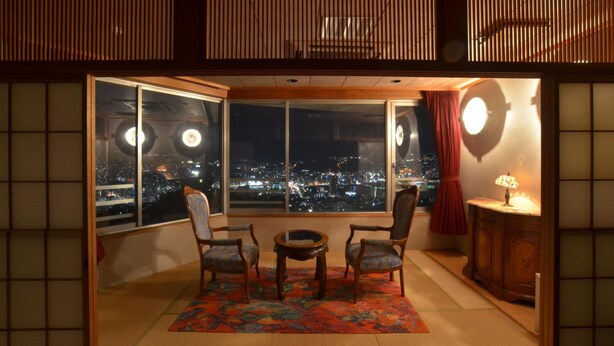 A traditional Japanese room with tatami flooring, a low table with chairs, a large window with a view of a cityscape at night, and a shoji screen. - Nagasaki Sky Hotel (Nagasaki)