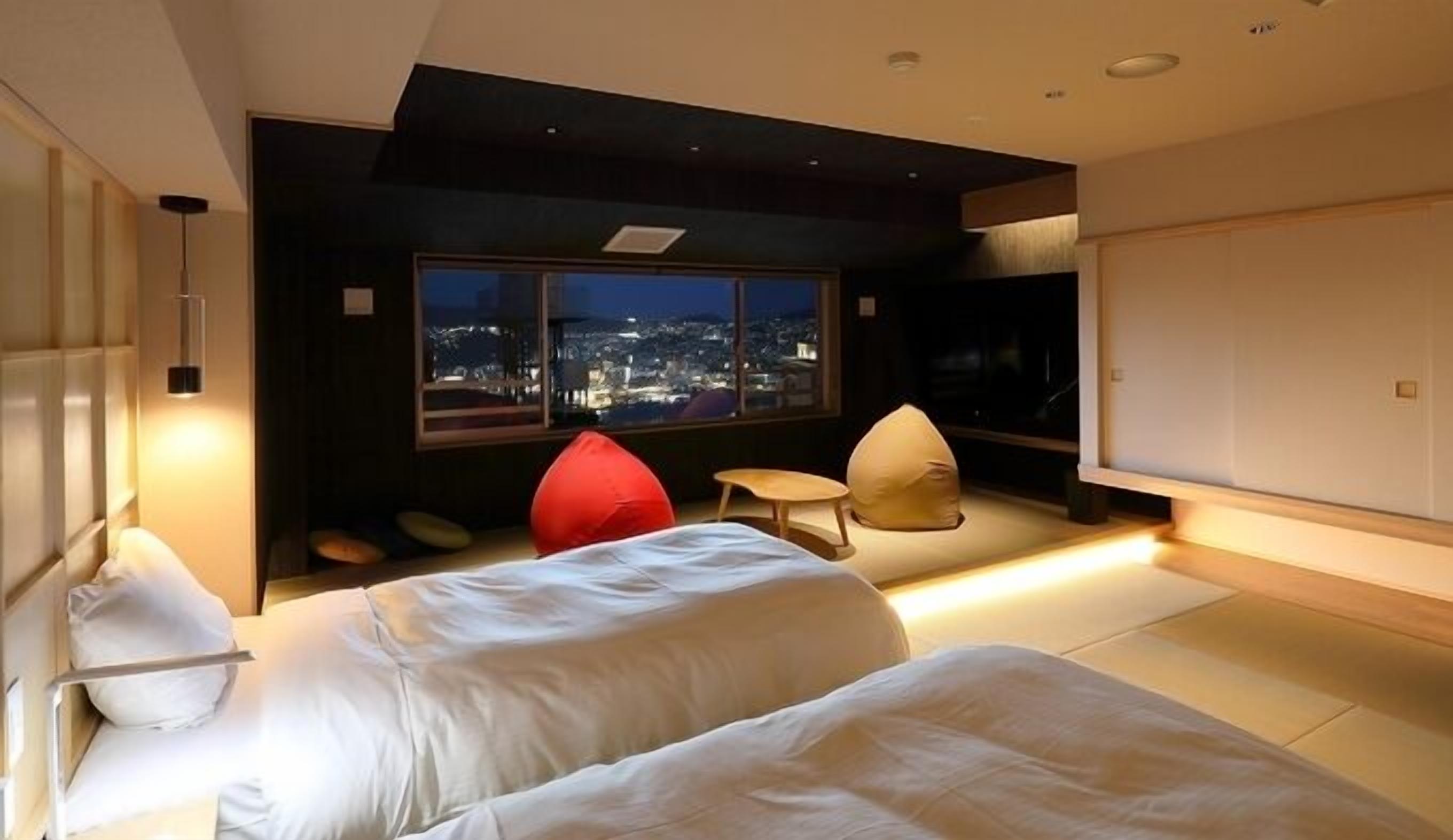 A modern hotel room with a city view, a bed, a red chair, and a small table.