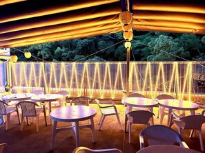 Een terras met ronde tafels en stoelen, overdekt door een luifel met lichtslingers.