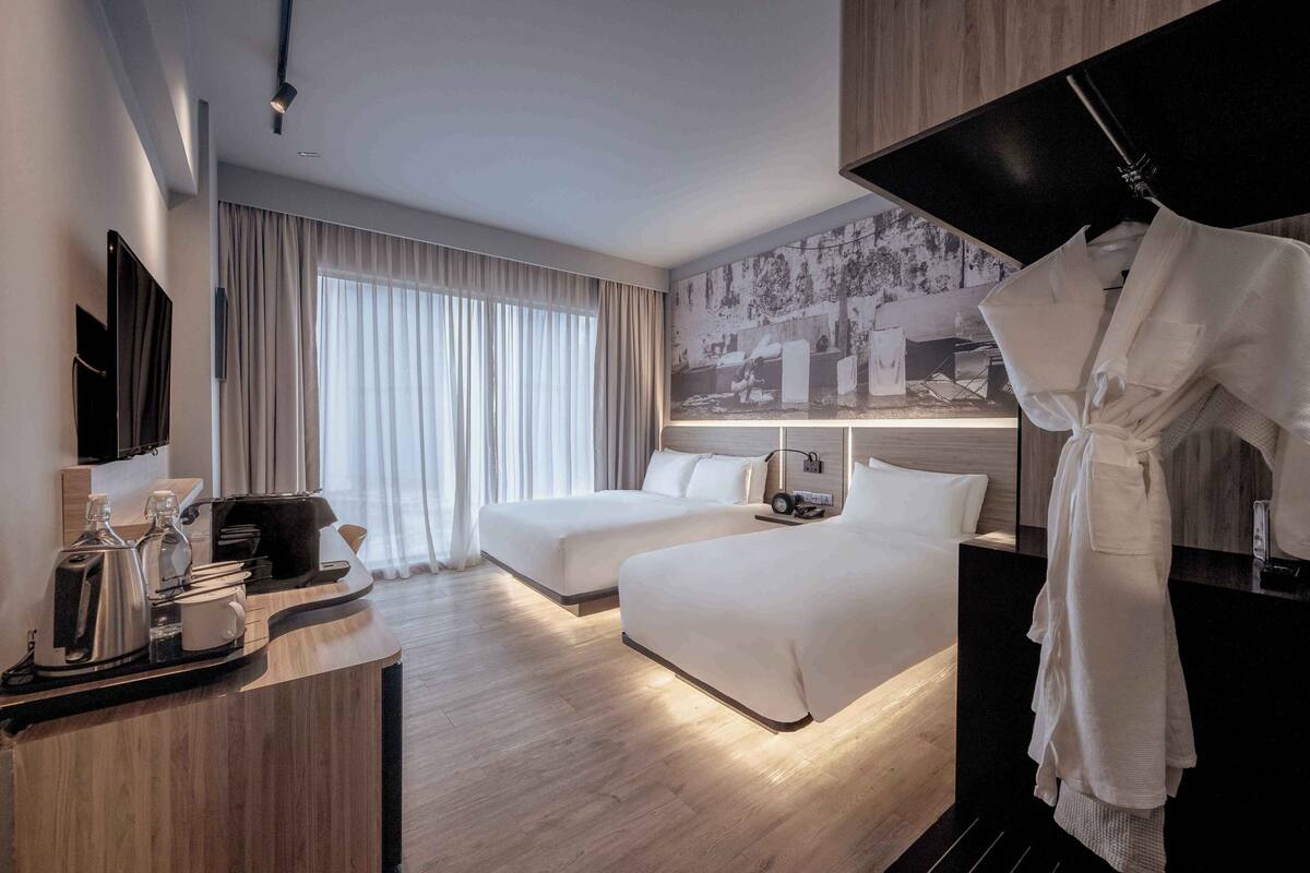 A hotel room with two beds, a desk with a coffee maker, a flat-screen TV, and a large window with curtains.