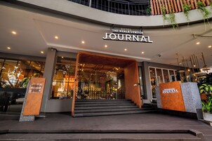 The entrance of The Kuala Lumpur Journal hotel with a modern design, featuring a glass facade, a staircase leading inside, and a signboard above the entrance.