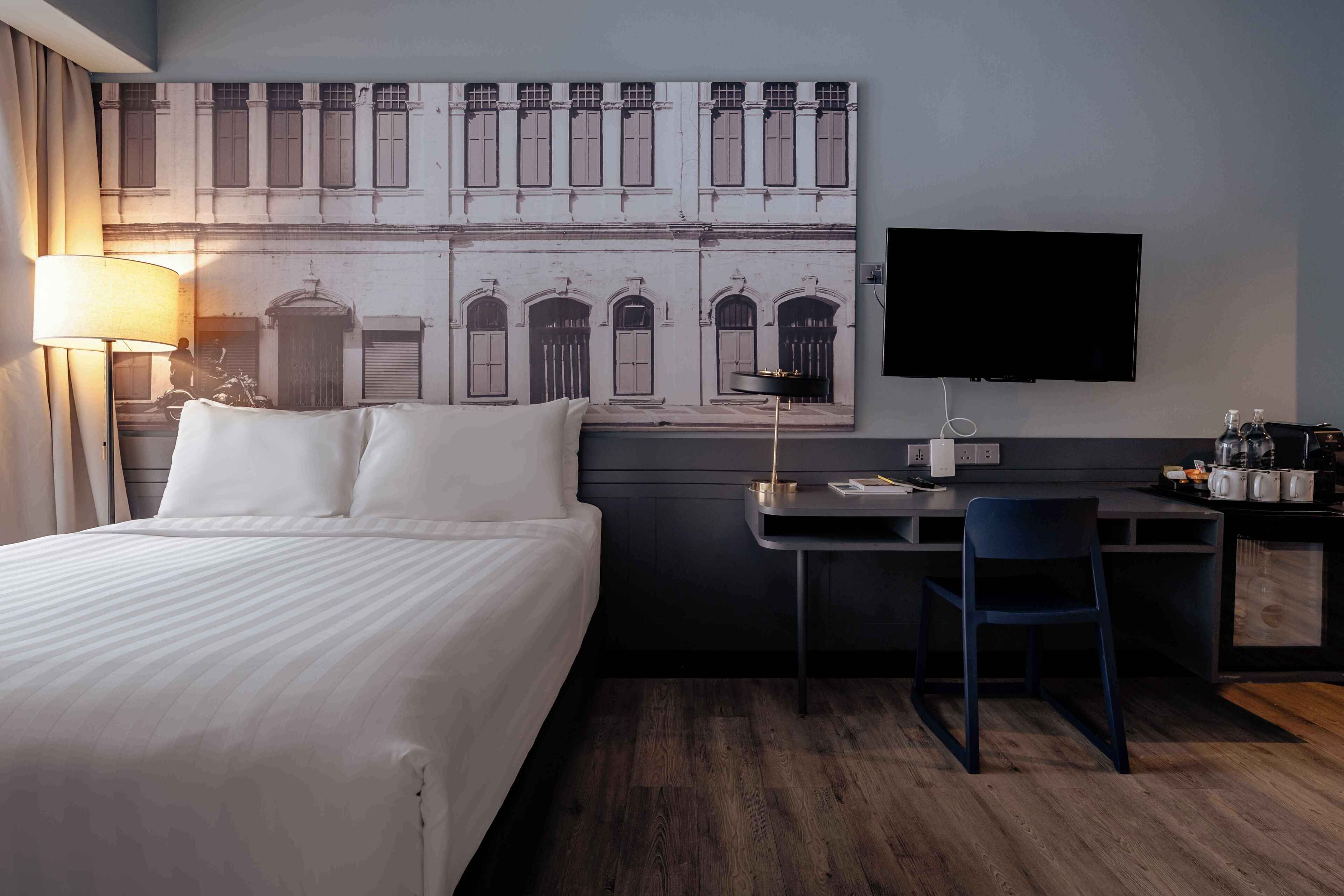 A hotel room with a large bed, a desk with a chair, a flat-screen TV, and a wall-mounted artwork depicting a building facade.
