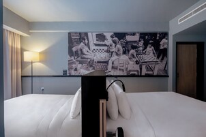 A hotel room with a large bed, a bedside table, a lamp, and a wall-mounted artwork depicting a chess game.