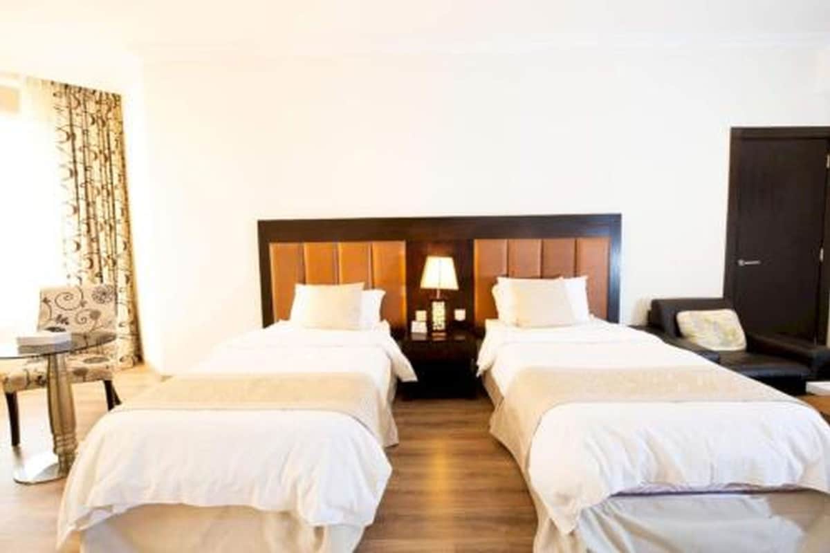 Habitación de hotel con dos camas individuales, cabecera de madera, mesita de noche con lámpara, una silla y un armario de color oscuro.