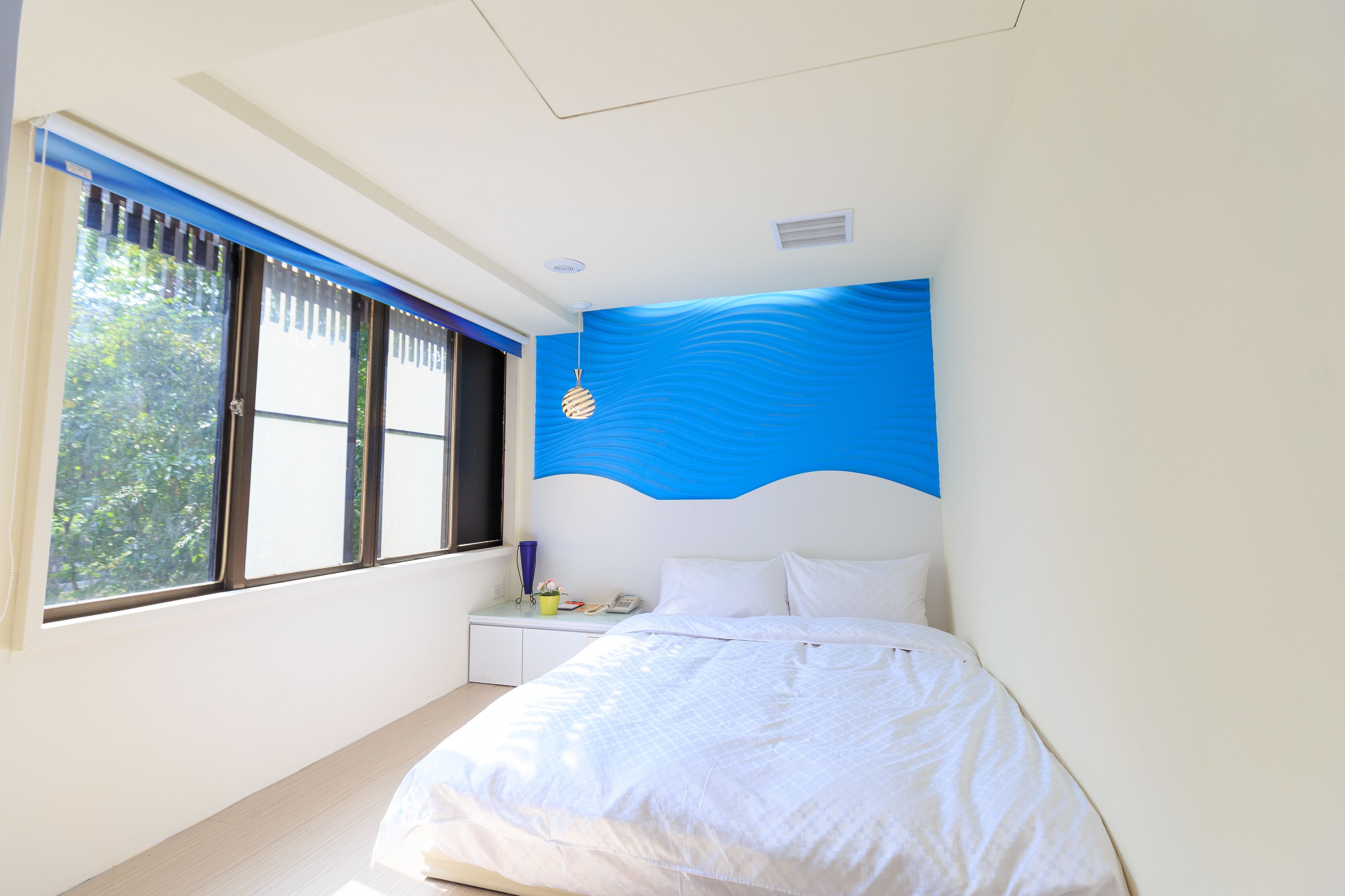Quarto com cama, mesa de cabeceira, janela com persianas e parede azul de destaque.