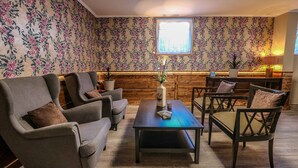 Una habitación acogedora con papel tapiz floral, un panel de madera decorativo y un ventanal que proporciona luz natural. Hay dos sillones y una mesa de centro de madera con un florero y una vela.