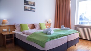 Una habitación de hotel con una cama, dos mesitas de noche, un ventanal con cortinas y un radiador.