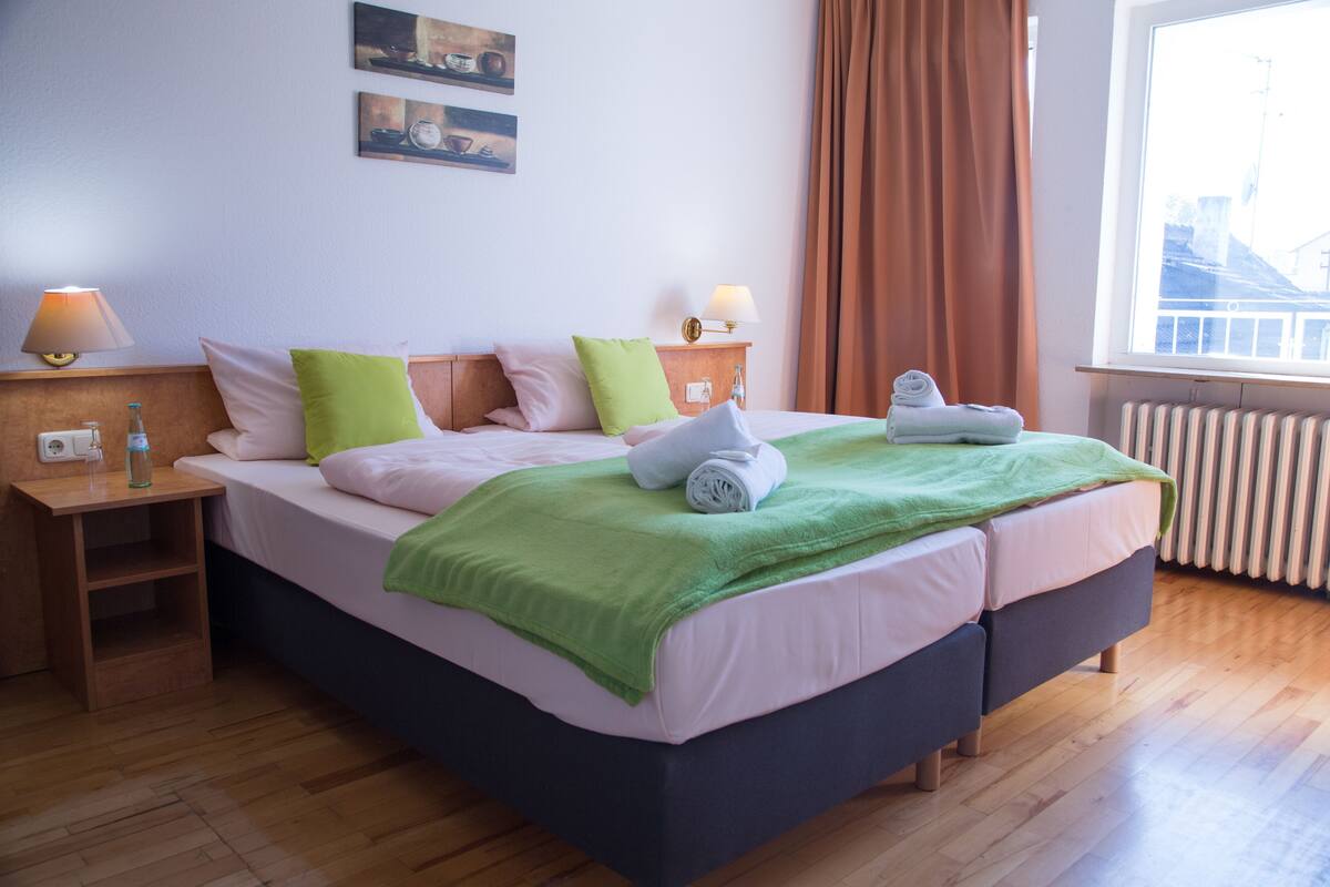 A hotel room with a bed, two bedside tables, a window with curtains, and a radiator.