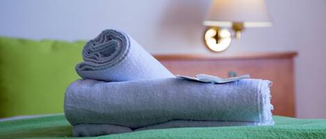 A neatly folded towel on a bed with a green blanket, a lamp on a bedside table, and a green pillow in a hotel room.