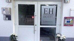 Entrada de hotel con puertas dobles, un letrero que dice "EH HOTEL" y dos plantas en macetas.