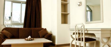 A room with a brown sofa, a white desk with a chair, a large mirror, and a window with curtains.