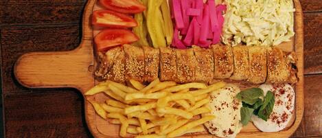 Una tabla de madera con diversos alimentos como rodajas de tomate, pepinillos, chucrut, un producto empanizado y frito, papas fritas y una salsa cremosa con guarnición.