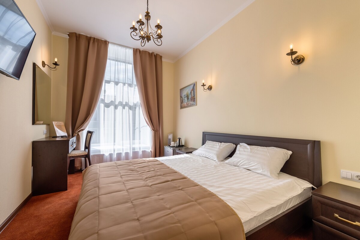A hotel room with a large bed, a desk, a chair, a desk lamp, a nightstand, a TV, and a chandelier.